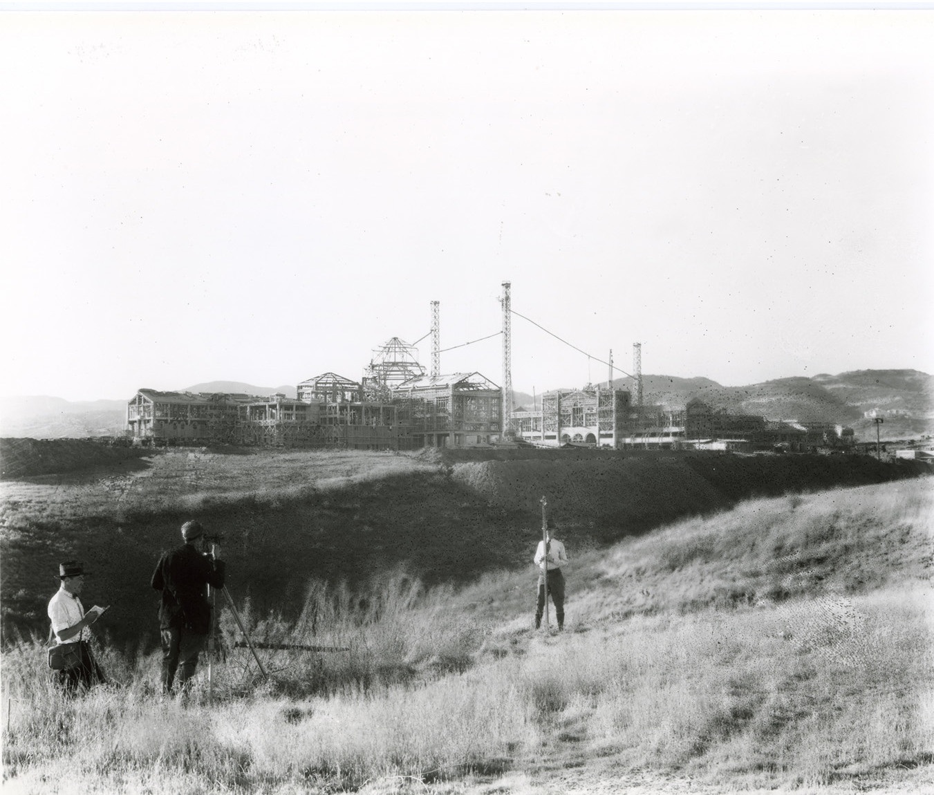 Black and white image of a field with construction in the background. There is a man standing in the foreground of the field with a pole, and two men further in the foreground - one is taking a picture of the first man, and the other is writing something down in a notebook.