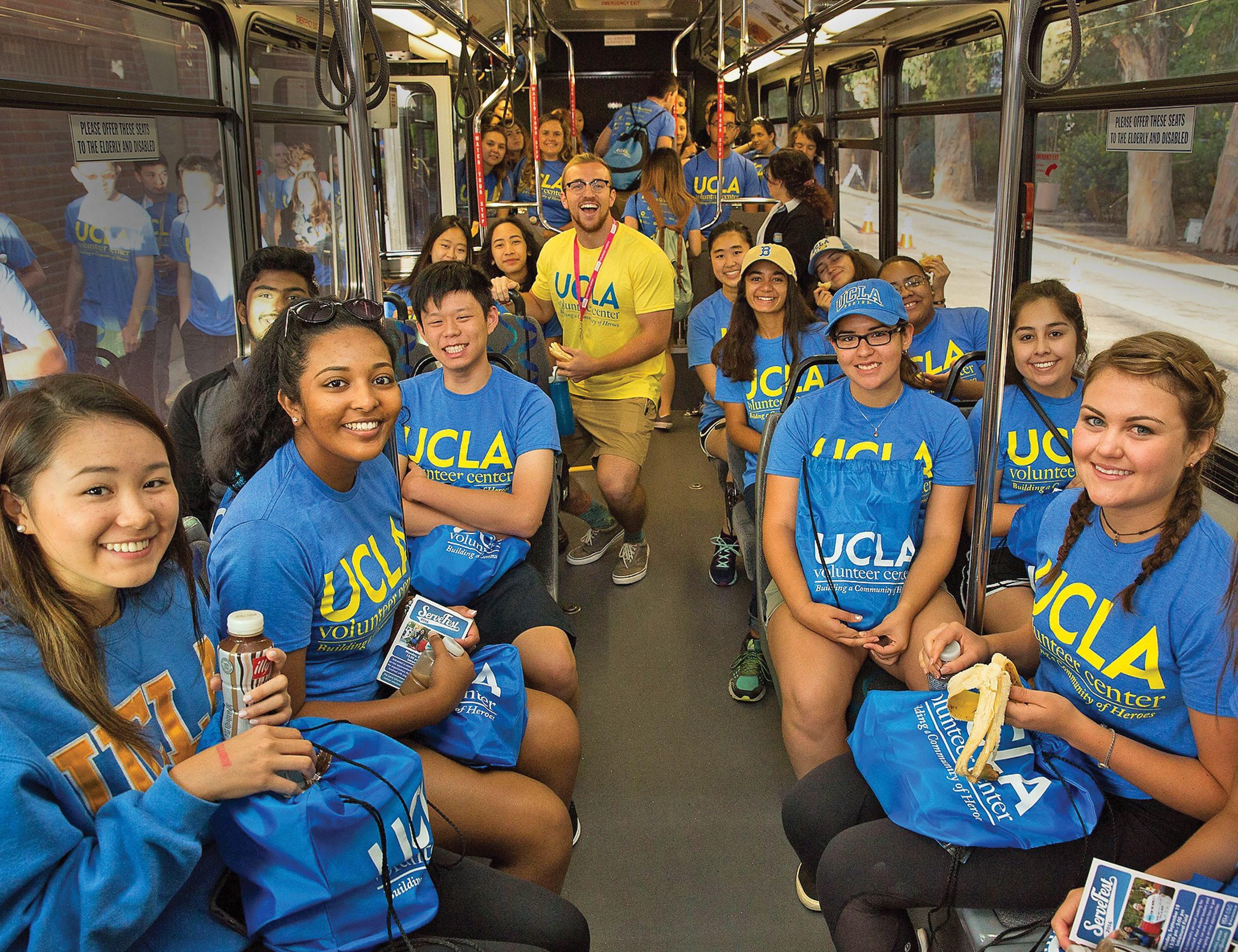 A group of UCLA students are wearing blue and yellow UCLA shirts on a bus. They are all smiling.